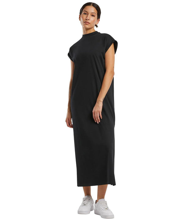 A person wears the Build Your Brand Women's long extended shoulder dress in black with white trainers, standing against a plain white background.