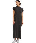 A woman wears the Build Your Brand women's vintage long dress, a loose-fitting, ankle-length dark grey piece with short sleeves and a mock neck. She pairs it with white trainers, her hair tied back and arms relaxed by her sides.