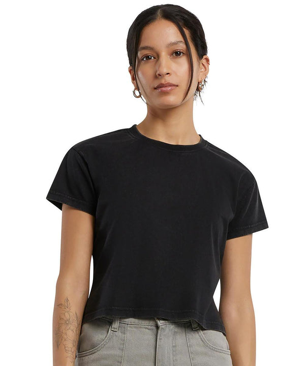 A person stands against a white background, wearing the Build Your Brand women's vintage cropped tee and grey high-waisted trousers. Their hair is tied back, with earrings and a tattoo on their left forearm.