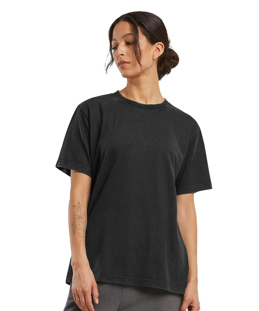 A person with dark hair pulled back is wearing the Build Your Brand Women's vintage everyday tee in black, paired with dark trousers, standing against a plain white background and looking down to their right.