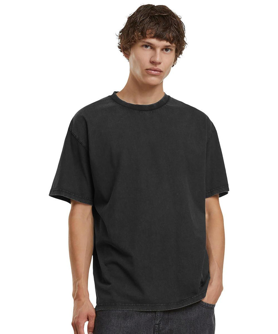 A young man with wavy brown hair wears the Build Your Brand Vintage heavy loose tee in black and dark trousers, standing against a plain white background with one hand in his pocket.