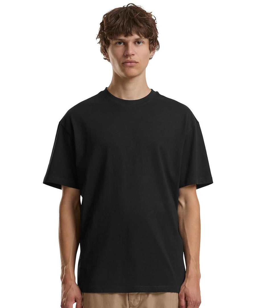 A person with short, wavy brown hair stands facing forwards, wearing the Build Your Brand Cotton loose tee in black and beige trousers against a white background.