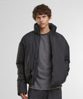 A person with short, wavy brown hair wears a Build Your Brand oversized puffer jacket over a grey shirt and dark trousers, standing against a plain white background with hands in the jacket pockets.