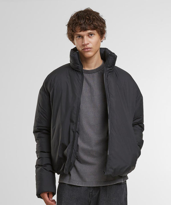A person with short, wavy brown hair wears a Build Your Brand oversized puffer jacket over a grey shirt and dark trousers, standing against a plain white background with hands in the jacket pockets.