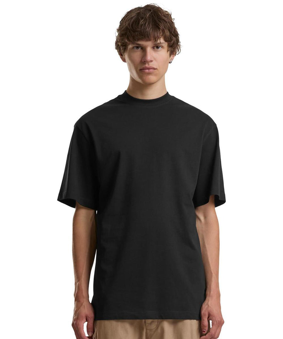 A person with short brown hair stands against a white background, wearing Build Your Brand’s Tall tee in plain black and beige trousers, facing forwards with a neutral expression.