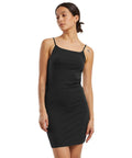A woman with her hair tied back and visible tattoos on her left arm and right shoulder wears the Build Your Brand Women's stretch jersey slim dress, a fitted, sleeveless black style with thin straps, against a white background.