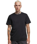 A person with short blond hair and visible neck and arm tattoos is wearing the Sorona regular tee by Build Your Brand with dark trousers, standing against a white background.
