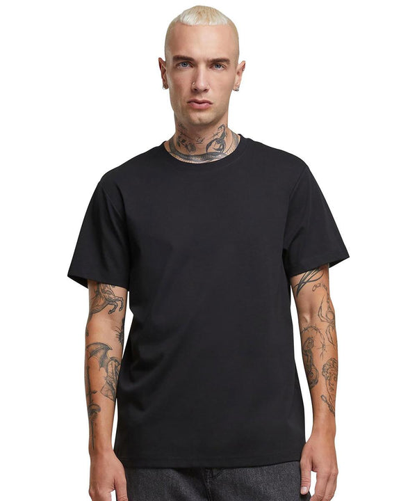 A person with short blond hair and visible neck and arm tattoos is wearing the Sorona regular tee by Build Your Brand with dark trousers, standing against a white background.