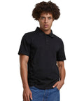 A person with curly hair is wearing the Sorona polo tee by Build Your Brand and blue jeans, standing against a plain white background.