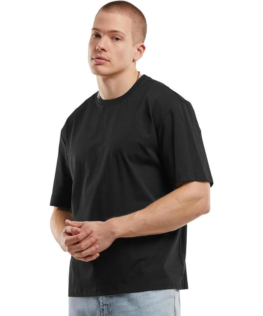 A man with short hair models the Sorona loose-fit tee by Build Your Brand in black, paired with light blue jeans. He stands against a white background, looking slightly to the side with his hands clasped.