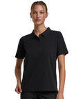 A woman faces forward wearing the Build Your Brand Women's Sorona polo tee in black, paired with black trousers. Her hair is tied back, her expression is neutral, and the background is plain white.