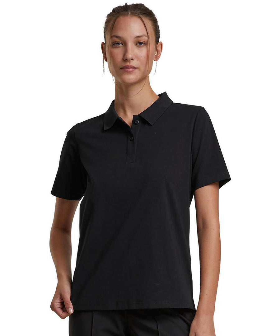 A woman faces forward wearing the Build Your Brand Women's Sorona polo tee in black, paired with black trousers. Her hair is tied back, her expression is neutral, and the background is plain white.