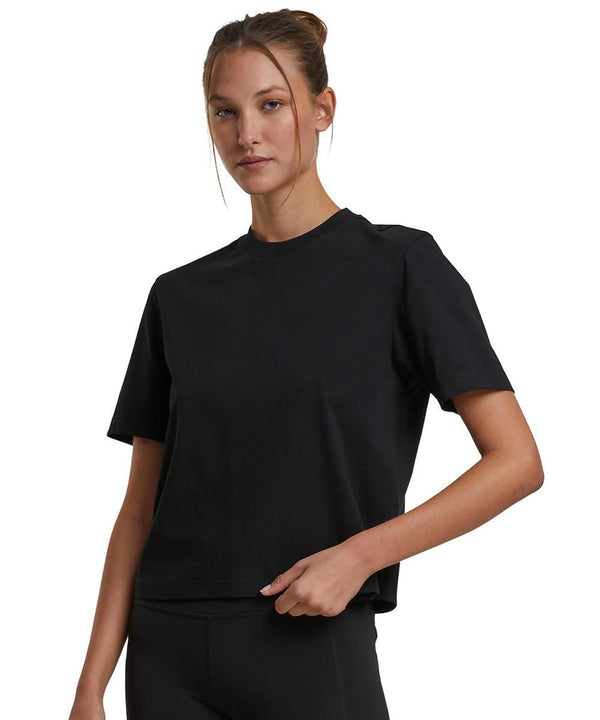A woman with light skin and brown hair in a bun wears the Build Your Brand Women's Sorona loose-fit tee in black with black trousers, standing against a white background and holding the hem of her shirt.