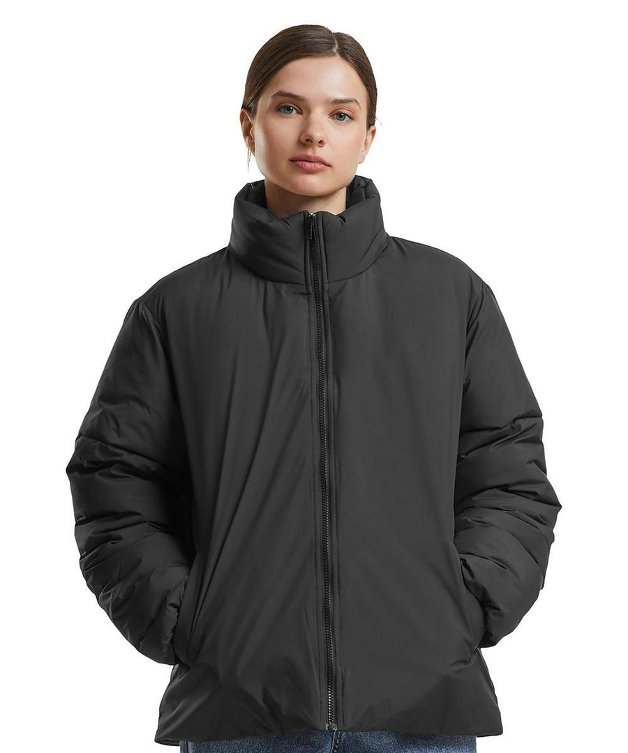 A person wears the Build Your Brand Women's loose puffer jacket in black, featuring a high collar and zip, hands in pockets, against a plain white background.