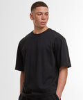 A person with short hair and freckles wears the Build Your Brand loose-fit tee in black and black trousers, standing with hands in pockets against a plain white background.