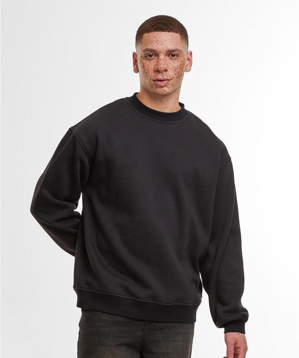 A person with short hair and freckles stands against a plain white background, wearing a Build Your Brand loose-fit sweatshirt in black and dark trousers, with one hand behind their back.