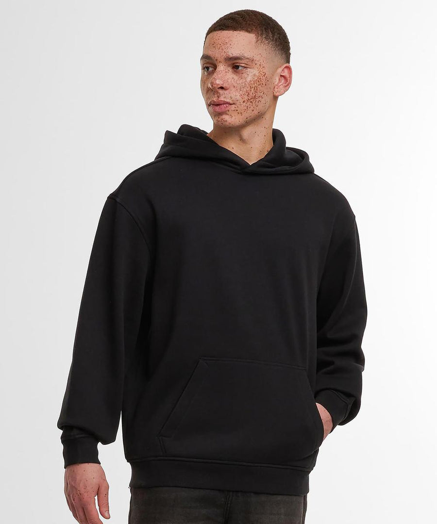 A person models the Build Your Brand loose-fit hoodie in black with dark jeans, standing against a white background and gazing to the side, one hand in the hoodie pocket.