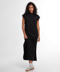 A person with curly hair stands against a plain white background wearing the Build Your Brand Women's Sorona long extended shoulder dress in black, paired with white trainers featuring light brown soles.