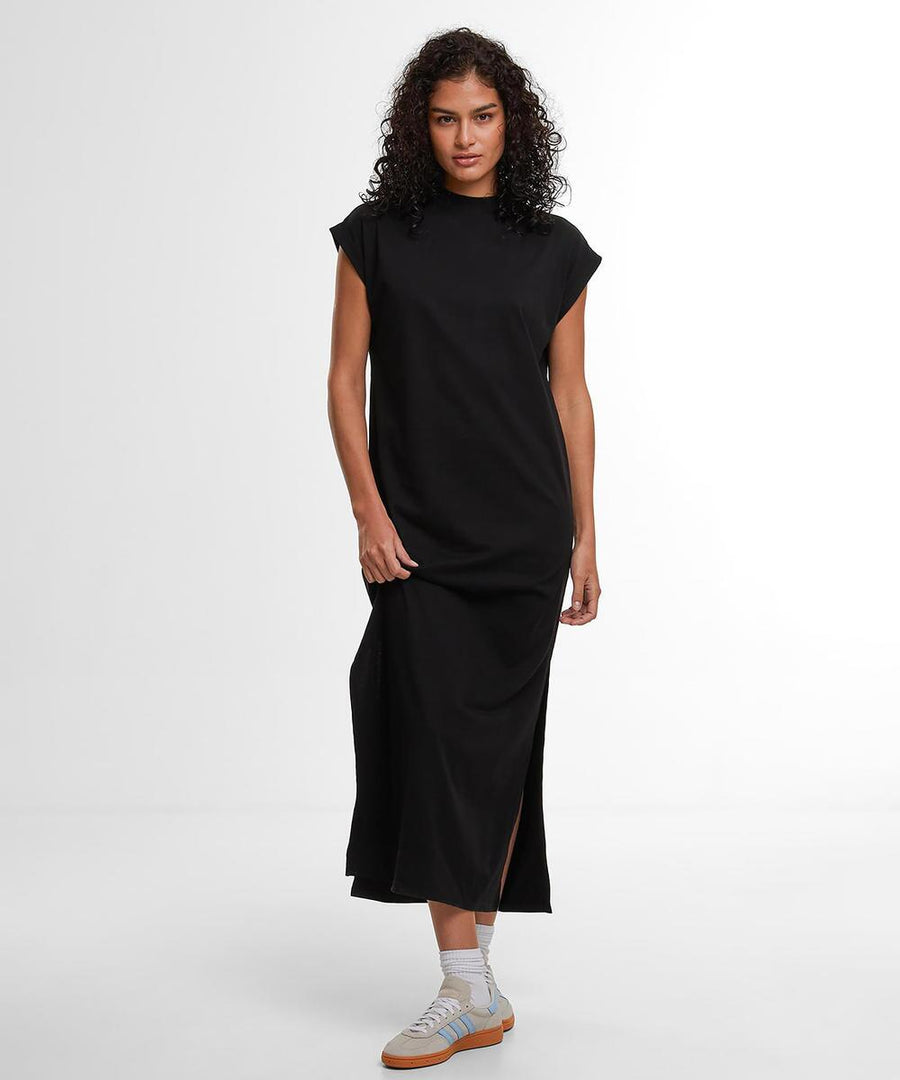 A person with curly hair stands against a plain white background wearing the Build Your Brand Women's Sorona long extended shoulder dress in black, paired with white trainers featuring light brown soles.