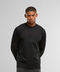 A person stands against a plain light grey background, wearing the Build Your Brand Sorona loose long sleeve shirt with dark trousers, hands behind their back and a neutral expression.