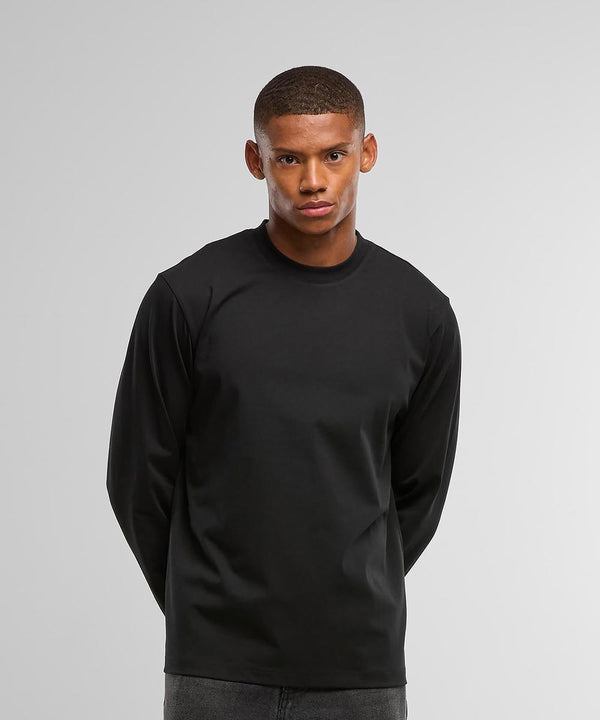 A person stands against a plain light grey background, wearing the Build Your Brand Sorona loose long sleeve shirt with dark trousers, hands behind their back and a neutral expression.