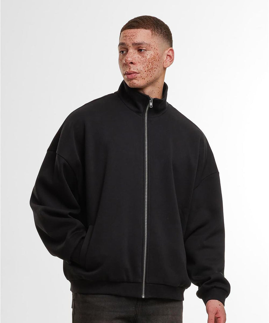 A person stands against a plain, light background wearing Build Your Brand's oversized zip jacket in black with a high collar and relaxed fit, paired with black trousers. Hands are in the jacket pockets.