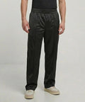 A person wearing a black T-shirt, Build Your Brand tricot track trousers in black with side seams, and white trainers stands against a plain light background.