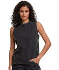 A person with curly hair wears the Build Your Brand Women's vintage washed sleeveless tee in faded black and matching black trousers, posing with one hand in their pocket against a plain white background.
