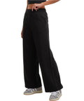 A person models Build Your Brand Women's wide-leg joggers in black, paired with a black cropped top and grey trainers with white stripes, set against a plain white background. The photo shows only the lower body and part of one arm.