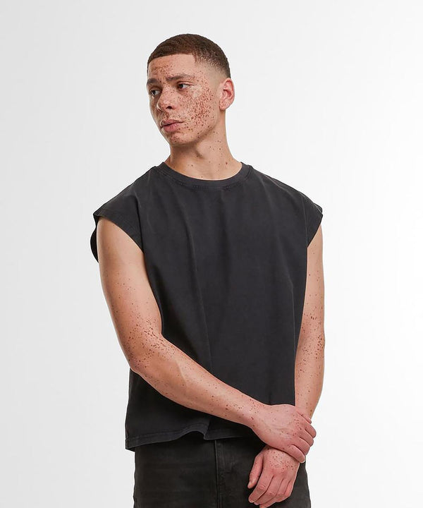 A person with short hair and visible freckles stands against a plain background, wearing the Build Your Brand Vintage cropped heavy sleeveless tee in black with dark trousers, arms crossed and gazing slightly to the side.