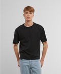 A young man with short light brown hair stands against a plain light grey background, wearing the Build Your Brand Cropped Oversize Tee in black and light blue jeans. He faces forward with his hands by his sides.