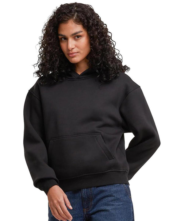 A woman with curly dark hair wears the Build Your Brand Women's loose-fit hoodie in black and blue jeans, standing against a plain white background.
