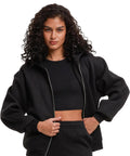 A person with curly dark hair wears the Build Your Brand Women's loose-fit zip hoodie in black over a crop top and trousers, standing with hands holding the hoodie, against a plain white background.