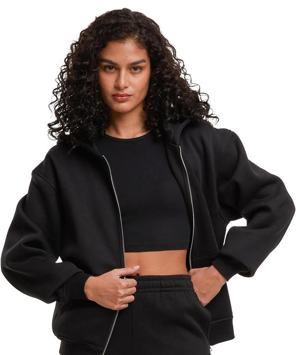 A person with curly dark hair wears the Build Your Brand Women's loose-fit zip hoodie in black over a crop top and trousers, standing with hands holding the hoodie, against a plain white background.