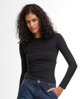 A person with curly dark hair wears the Build Your Brand Women’s Ultra Slim Long Sleeve black top and blue jeans, standing against a white background with one hand in a pocket.