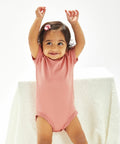 A toddler in a pink Babybugz BZ010 with a bow in their hair smiles with arms raised, standing against a white background.