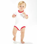 A toddler with blond hair is standing and walking towards the camera, wearing a white Babybugz BZ019 onesie with red trim, holding a small object in one hand.