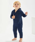 A young child stands barefoot, wearing a navy blue Babybugz BZ025 hooded onesie with front pockets, looking slightly upward.