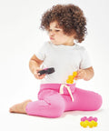 A toddler with curly hair wears a white BZ033 shirt from Babybugz and pink pants, sitting on the floor and holding a toy in each hand.
