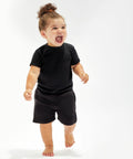 A toddler wearing a Babybugz BZ061 black t-shirt and black shorts stands barefoot, smiling with an open mouth against a plain white background.