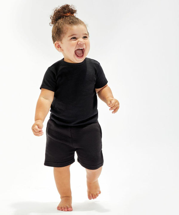 A toddler wearing a Babybugz BZ061 black t-shirt and black shorts stands barefoot, smiling with an open mouth against a plain white background.