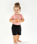 A toddler with curly hair stands barefoot wearing a pink shirt and black shorts, holding a small orange Babybugz BZ065.