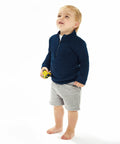 A toddler wearing a navy sweater and gray shorts stands barefoot, holding a yellow Babybugz BZ066 toy car, looking upwards with a curious expression.