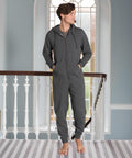A person stands on a staircase landing in front of a tall window, wearing a dark gray CC001 zippered onesie with a hood by Comfy Co.