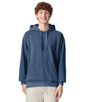 A young person with curly hair is smiling at the camera whilst wearing a blue Comfort Colours hooded sweatshirt and light trousers against a plain white background.