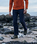 A person in a Craghoppers CR233 jacket and blue pants walks on a rocky, seaweed-covered beach with the ocean in the background.