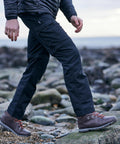 A person walking on a rocky shoreline wearing dark pants, a quilted jacket, and Craghoppers CR235 brown hiking boots with orange laces.