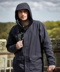 A person stands outdoors wearing a dark Craghoppers CR305 rain jacket with a hood. They have a backpack strap over one shoulder and are in front of a blurred background of trees and a stone structure.