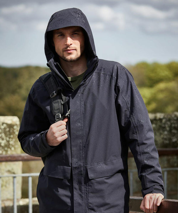 A person stands outdoors wearing a dark Craghoppers CR305 rain jacket with a hood. They have a backpack strap over one shoulder and are in front of a blurred background of trees and a stone structure.
