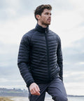 A man wearing a Craghoppers CR307 quilted jacket and gray pants is walking outdoors near a coastline under a cloudy sky.